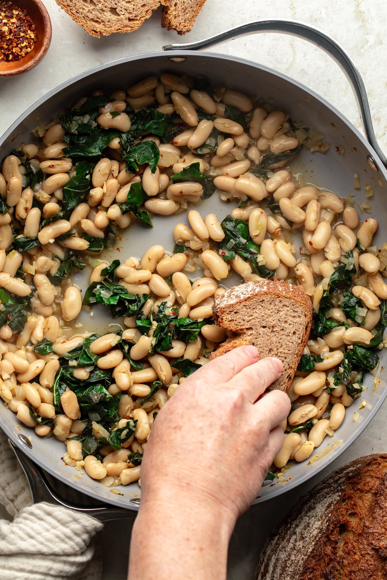 overhead view of a hand dragging a piece of bread through a skillet full of braised white beans and kale.