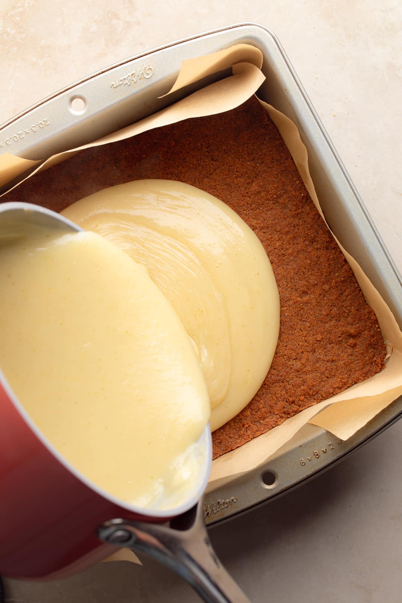 Overhead view of liquid key lime pie filling being poured over a brown cookie crust in a baking pan.
