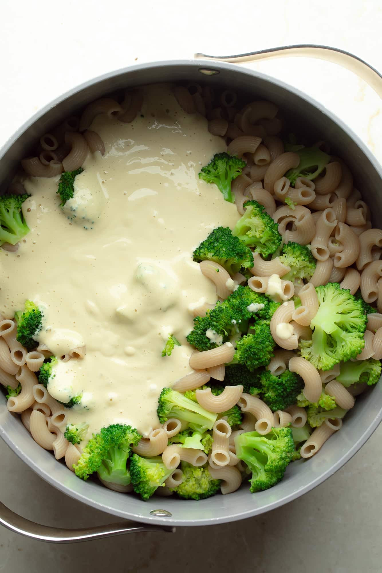 overhead view of white vegan cheese sauce on top of cooked broccoli and pasta in a large pot.
