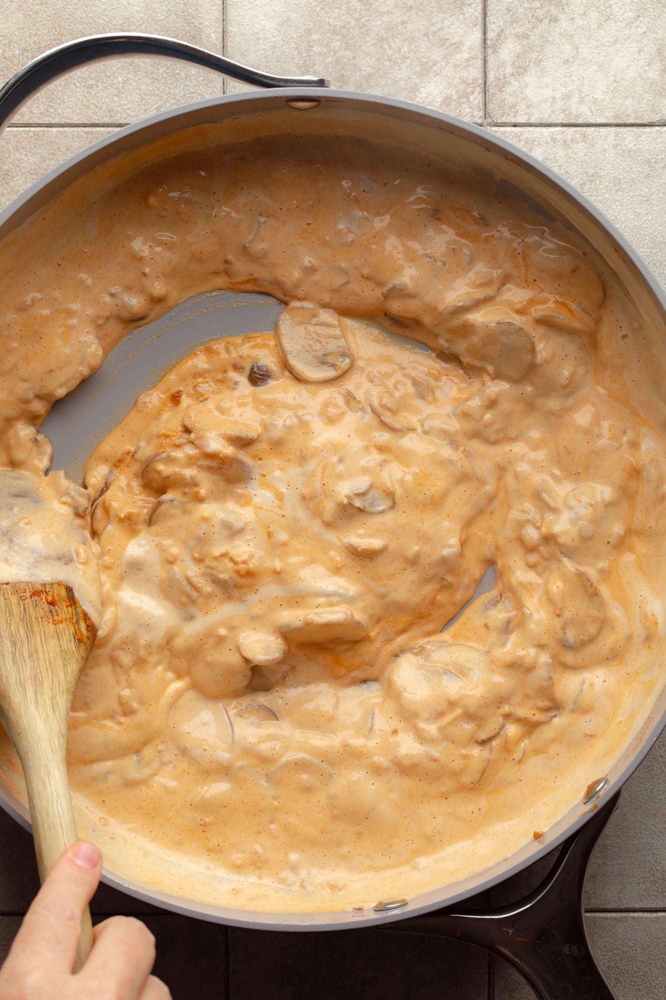 overhead view of a hand using a wooden spoon to stir a creamy gochujang sauce in a large skillet.