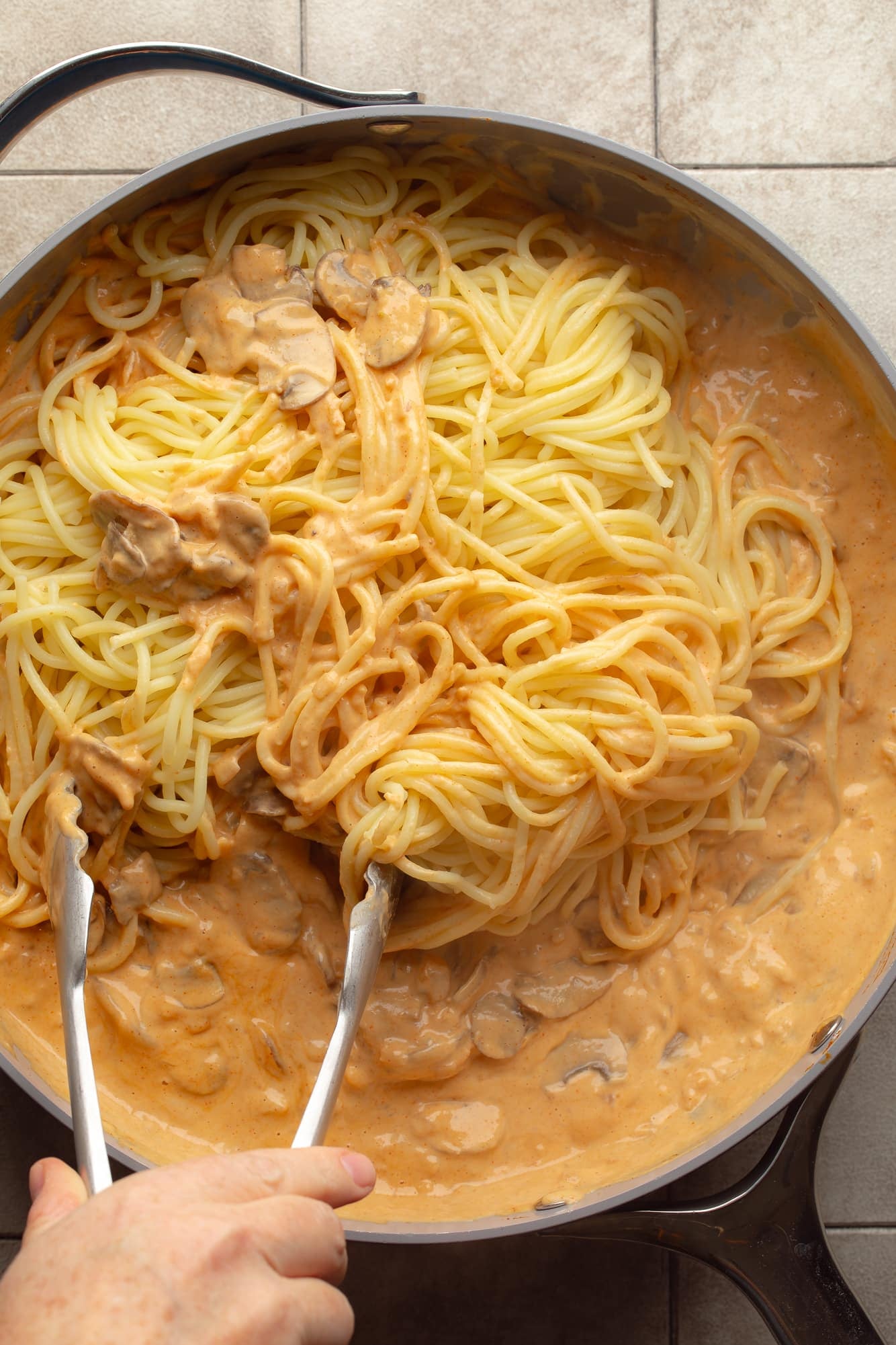 overhead view of a hand using metal tongs to toss noodles in a creamy gochujang sauce in a skillet.