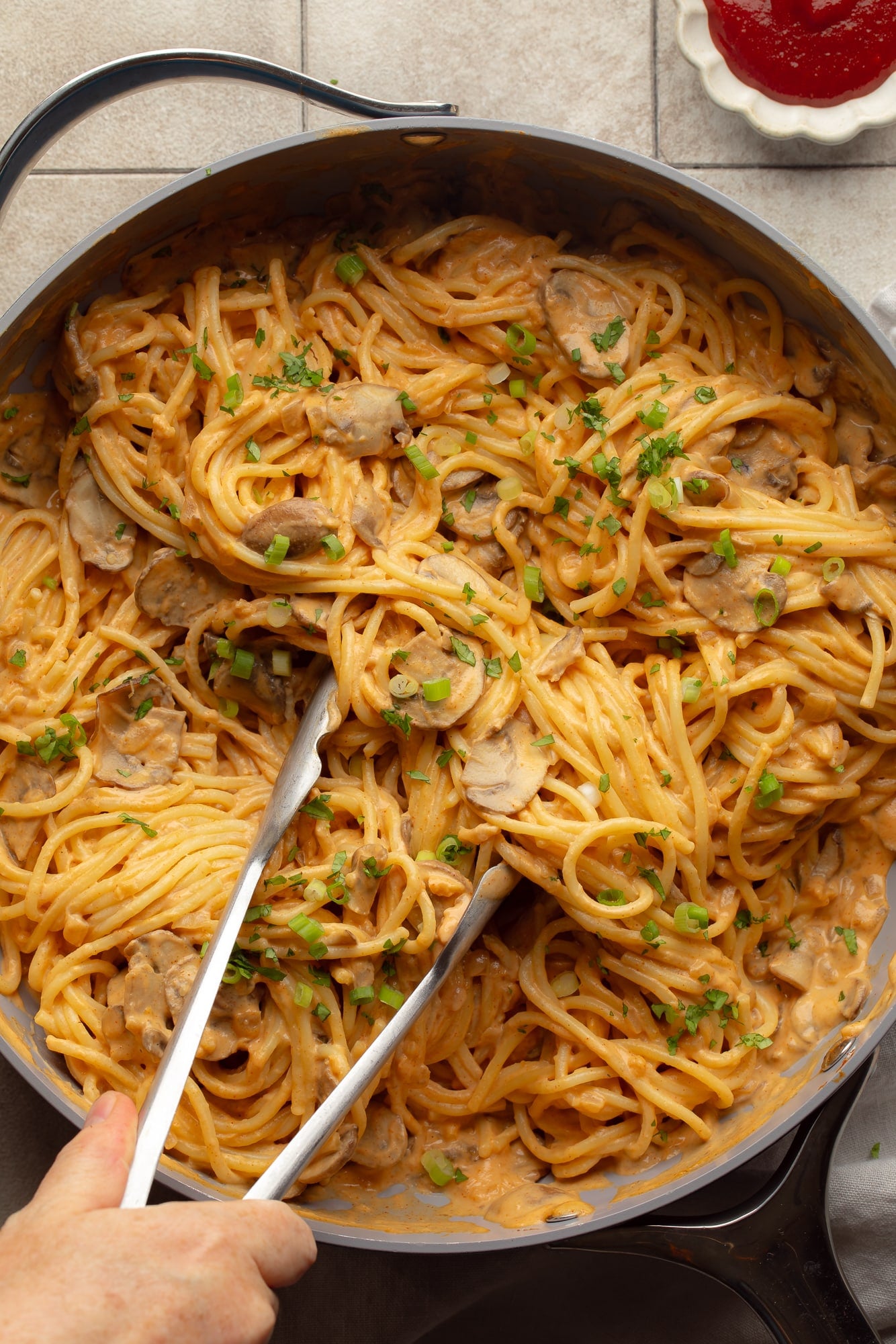 overhead view of a hand holding metal tongs in a skillet full of creamy gochujang noodles.