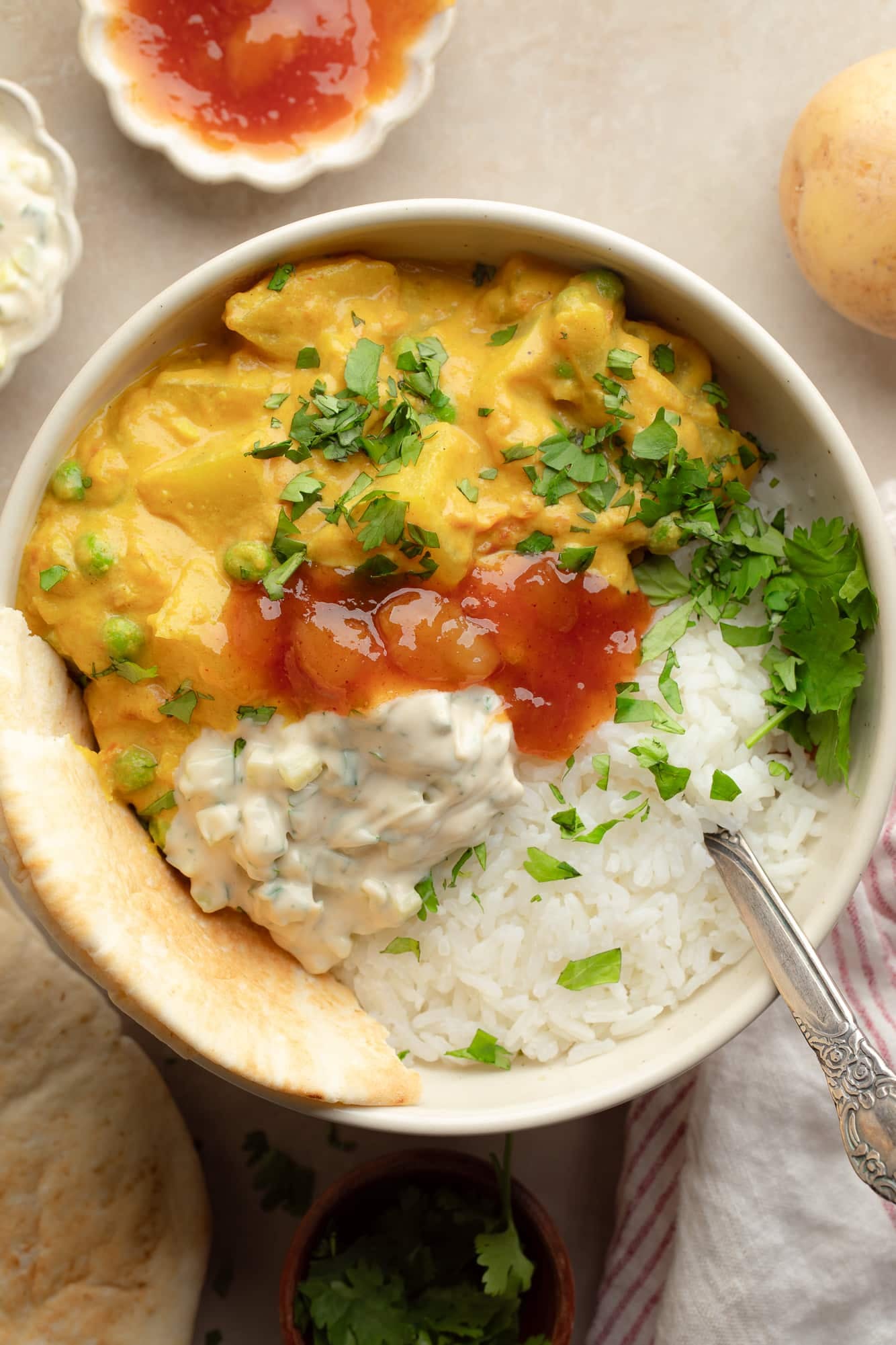 overhead view of a bowl of potato curry topped with cilantro, raita, and mango chutney.