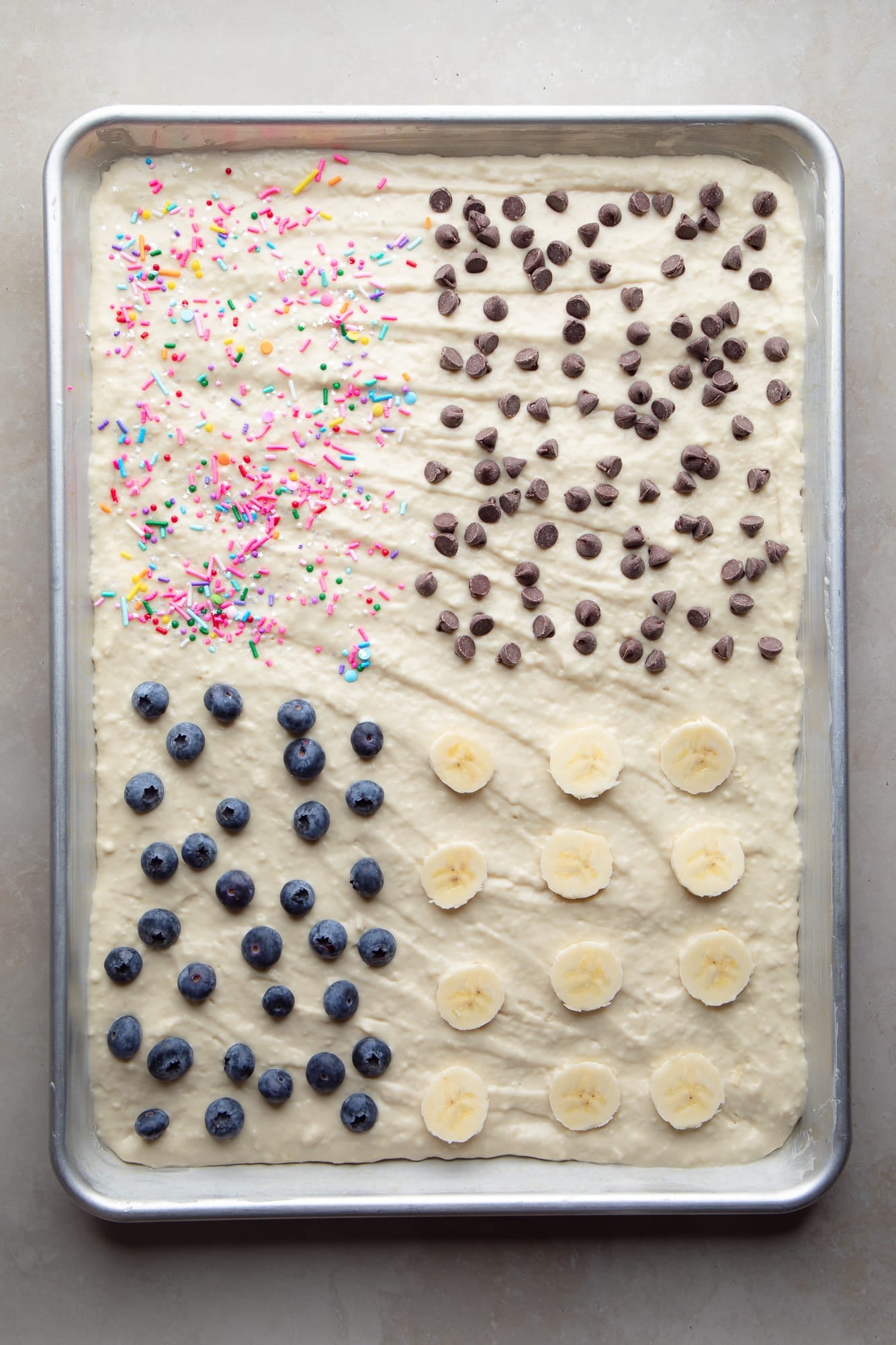 overhead view of uncooked sheet pan pancakes topped with sprinkles, fruit, and chocolate chips on a metal pan.