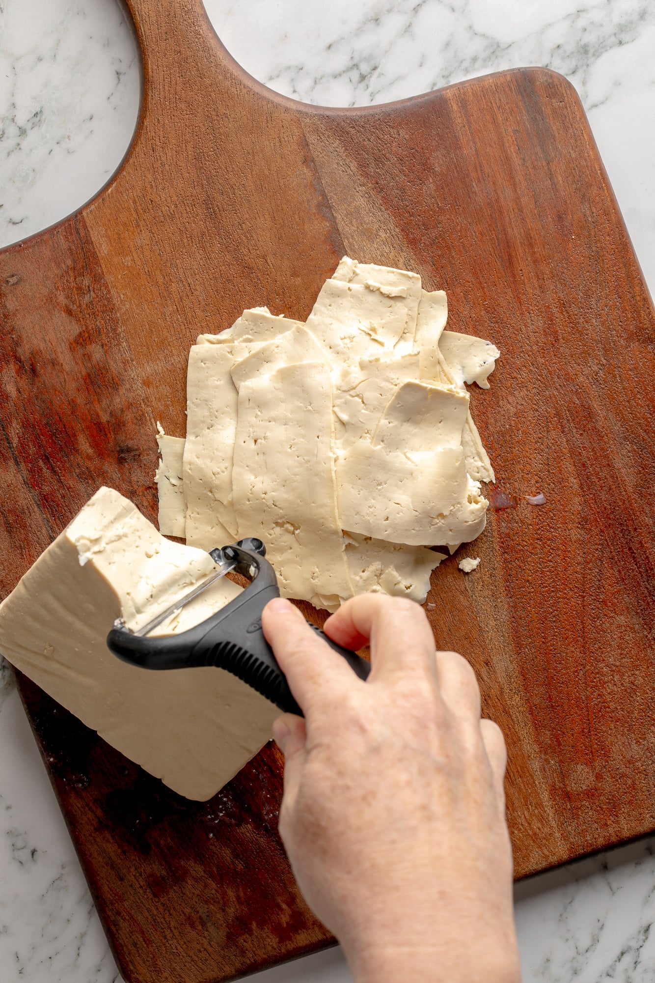overhead view of a hand shaving a block of tofu with a vegetable peeler.
