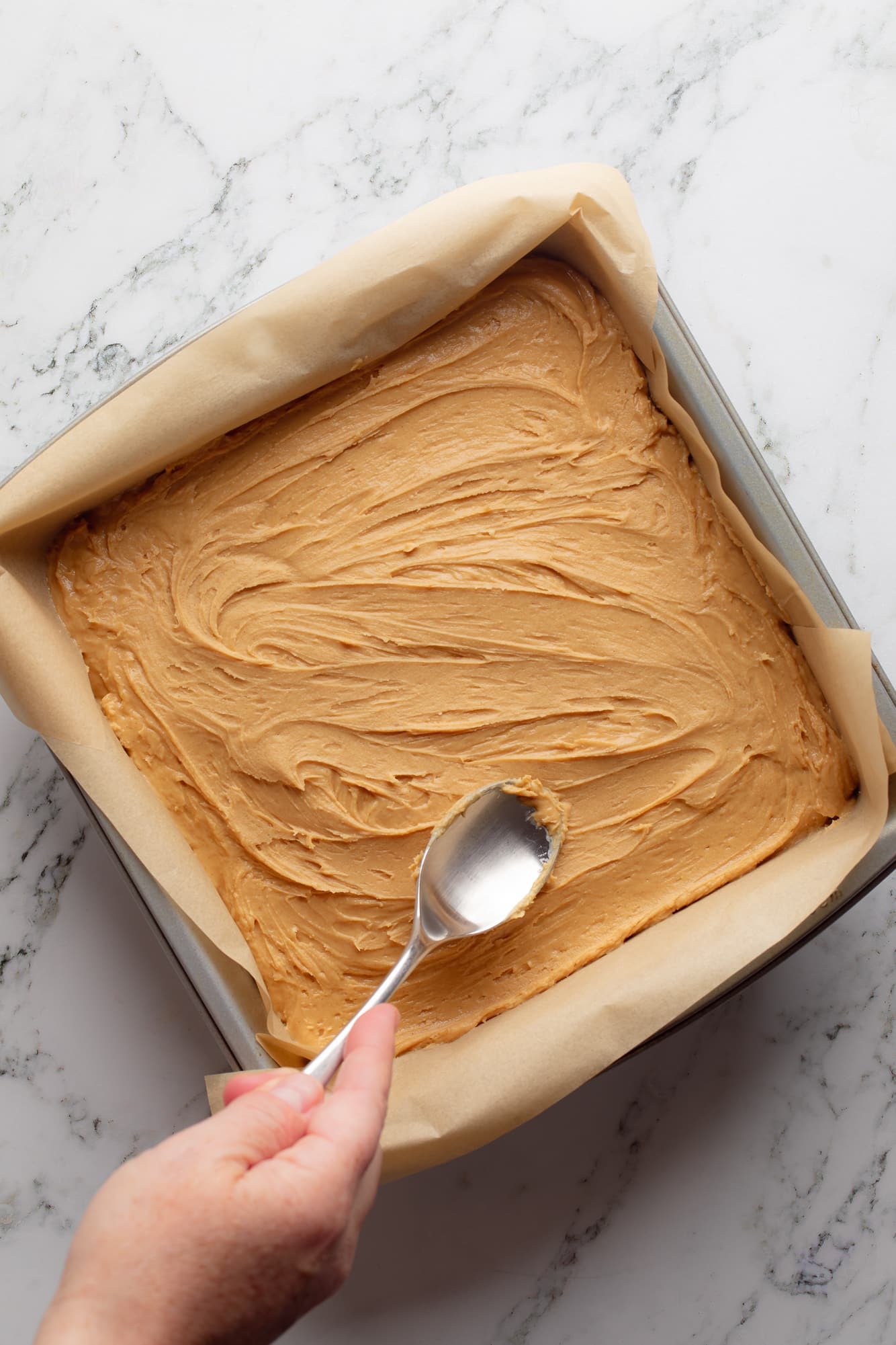 overhead view of a hand using a spoon to spread the peanut butter layer for Chocolate Peanut Butter Oatmeal Bars in a baking pan.