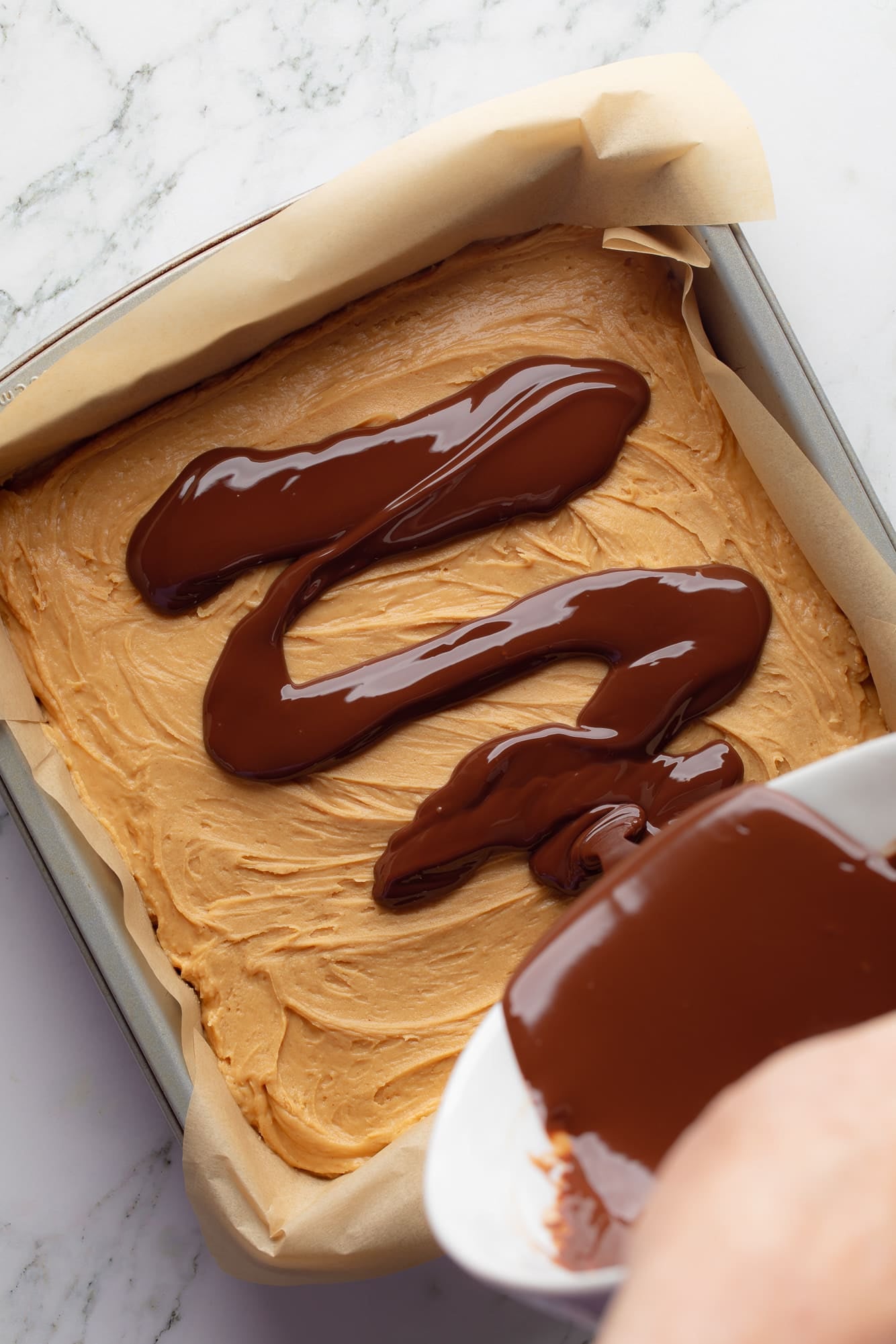 overhead view of a hand pouring melted chocolate from a bowl over the peanut butter layer of Chocolate Peanut Butter Oatmeal Bars in a baking pan.