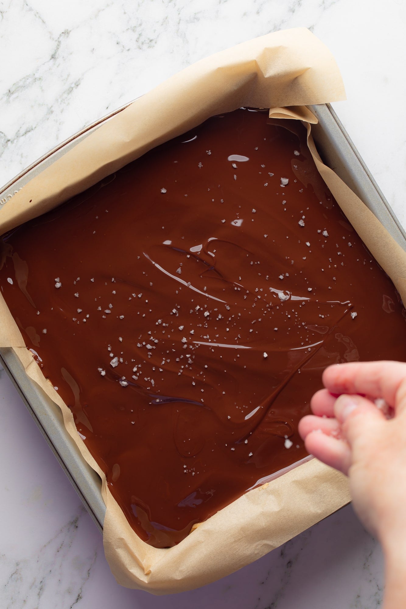 overhead view of a hand sprinkling salt on top of the chocolate layer of Chocolate Peanut Butter Oatmeal Bars in a baking pan.