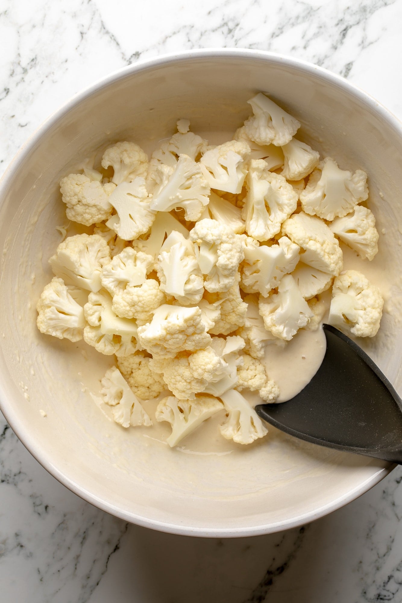 overhead view of cauliflower florets being tossed in a batter in a large white bowl.