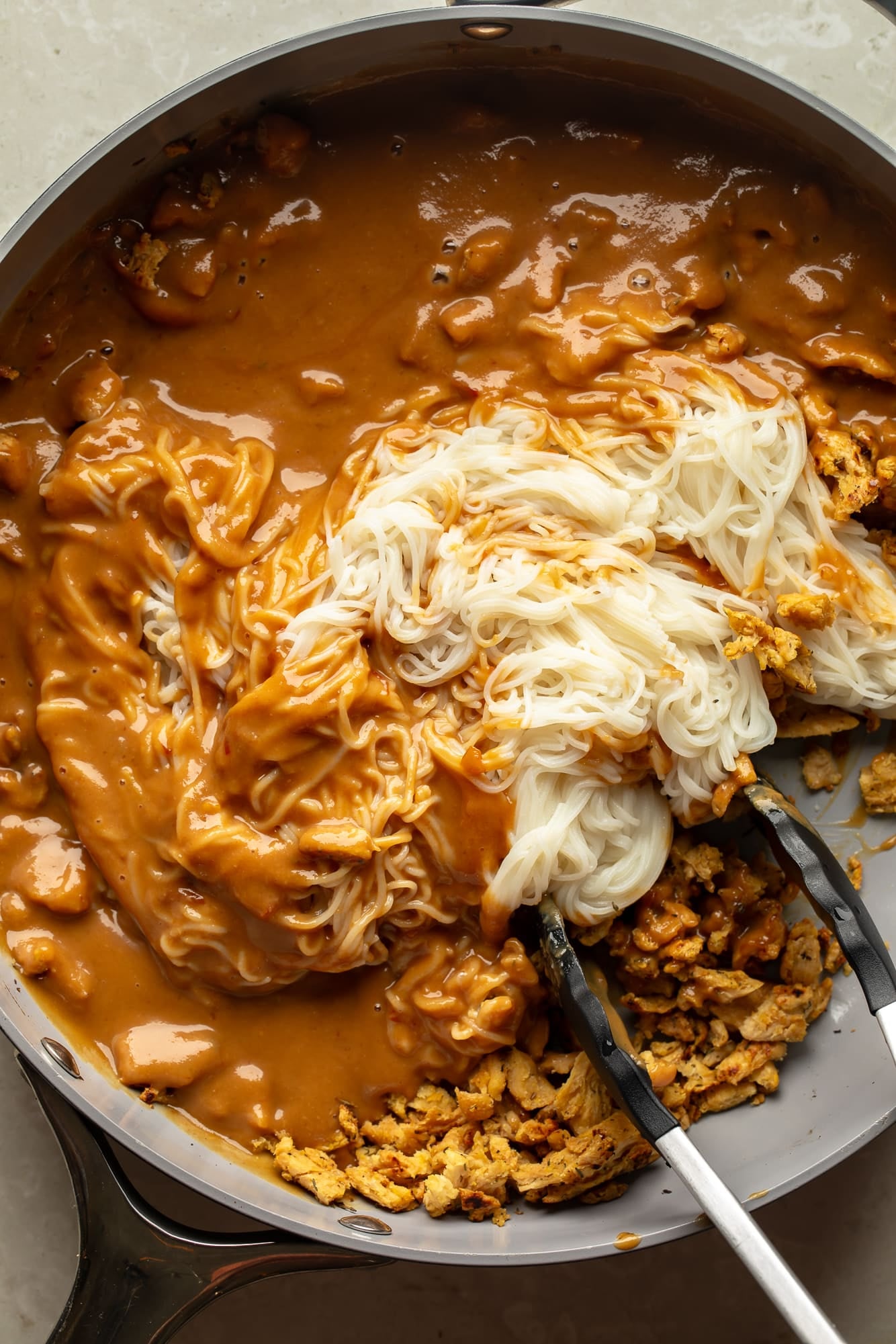 overhead view of tongs stirring noodles, soy curls, and peanut sauce together in a skillet for spicy peanut vegan "chicken" noodles.