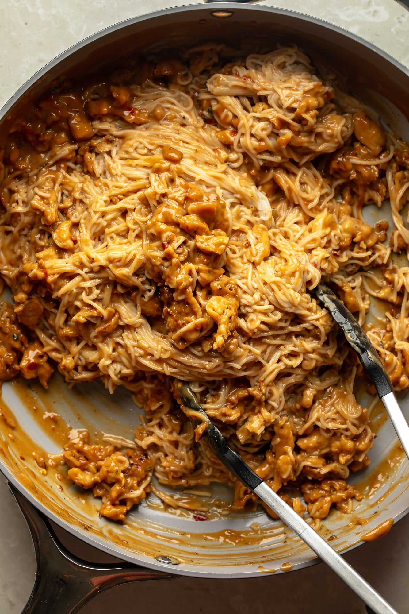 overhead view of tongs stirring noodles, soy curls, and peanut sauce together in a skillet for spicy peanut vegan "chicken" noodles.