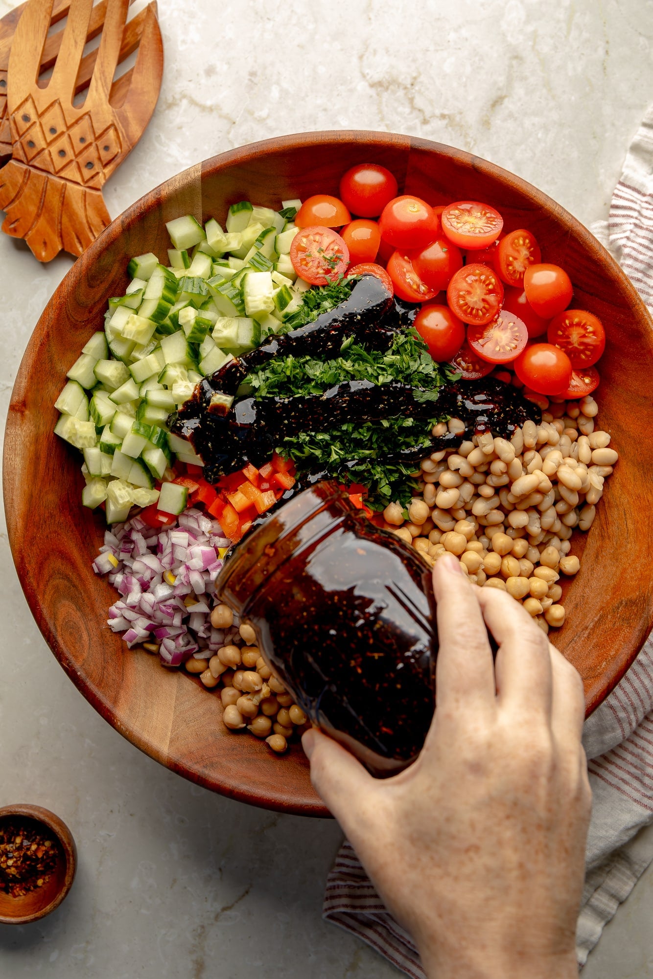 overhead view of a hand pouring balsamic dressing over the ingredients for a vegan dense bean salad in a large bamboo bowl.