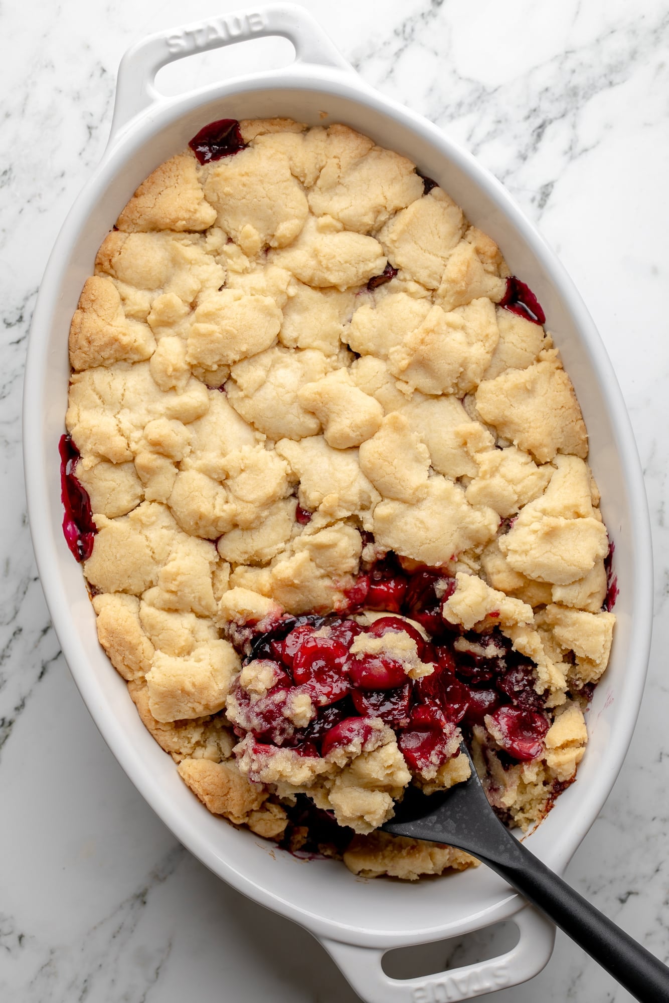overhead view of a black spoon lifting a scoop of vegan cherry cobbler from a large baking dish.