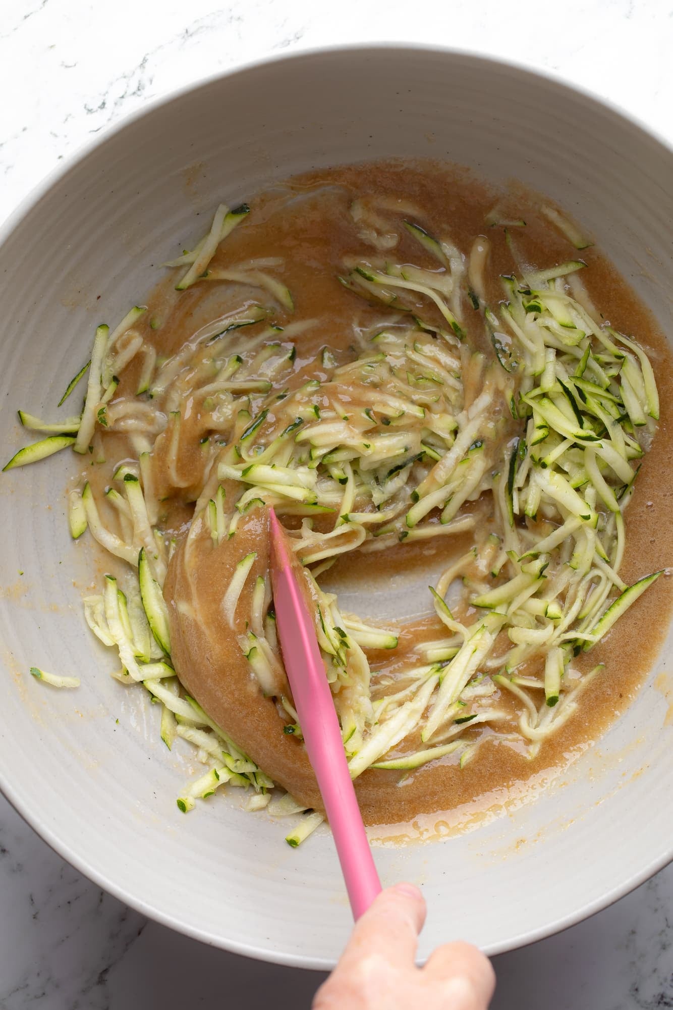 overhead view of a pink spatula stirring grated zucchini into vegan zucchini muffin batter.