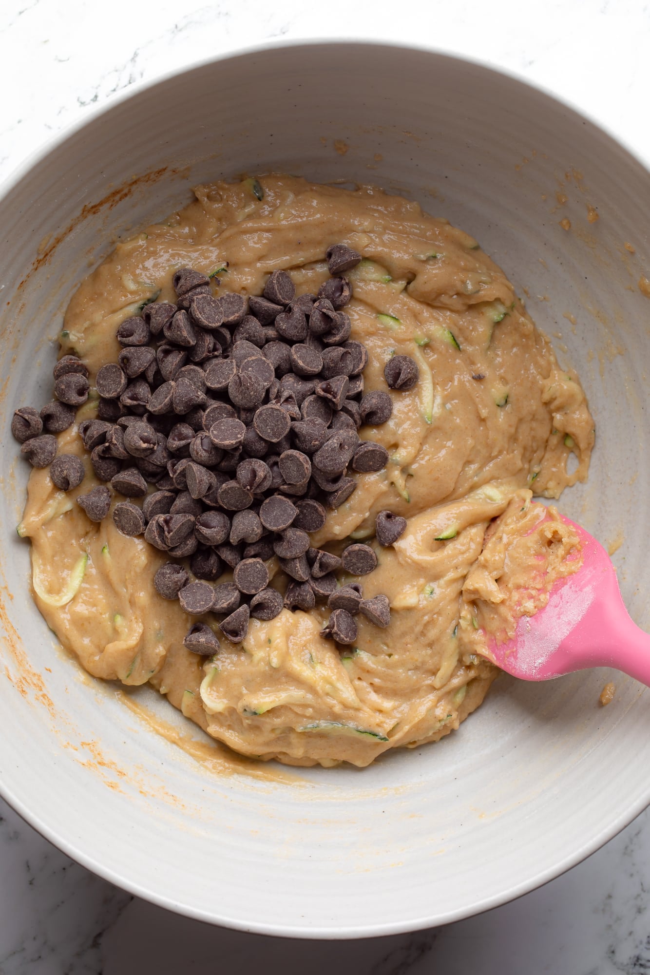 overhead view of a pink spatula stirring chocolate chips into vegan zucchini muffin batter.