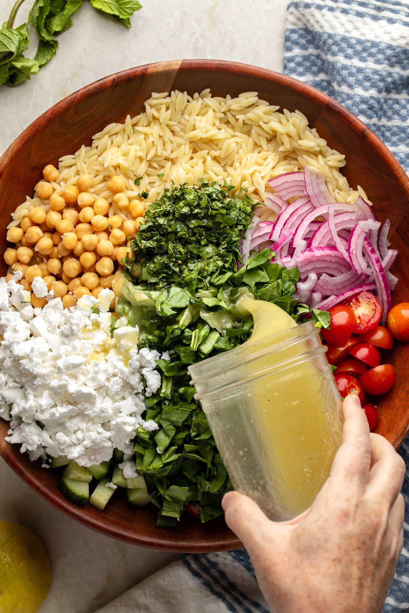 overhead view of a jar of lemon dressing being poured over a lemon orzo pasta salad in a large bamboo bowl.
