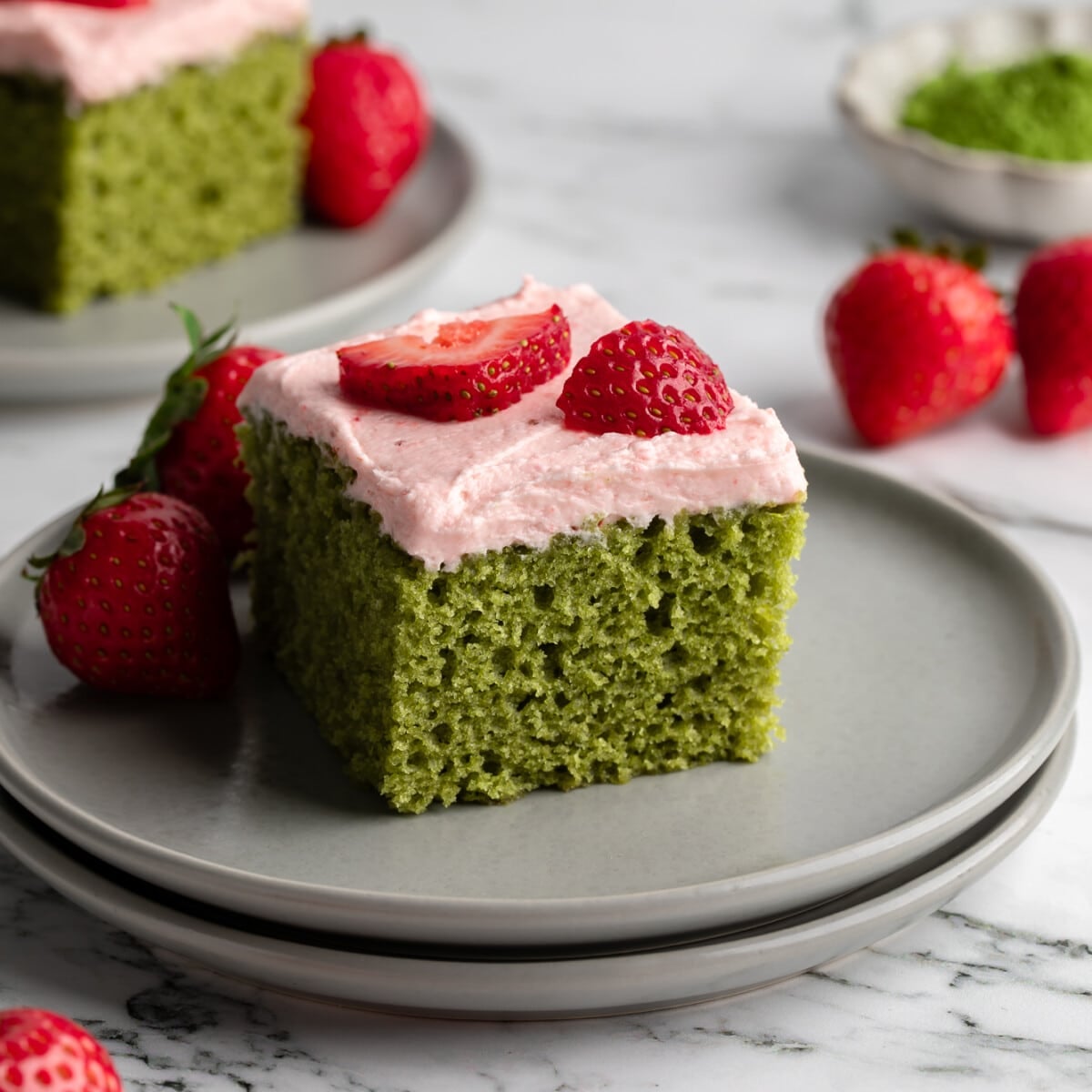 square image of a slice of vegan strawberry matcha cake topped with fresh strawberries on a grey plate.