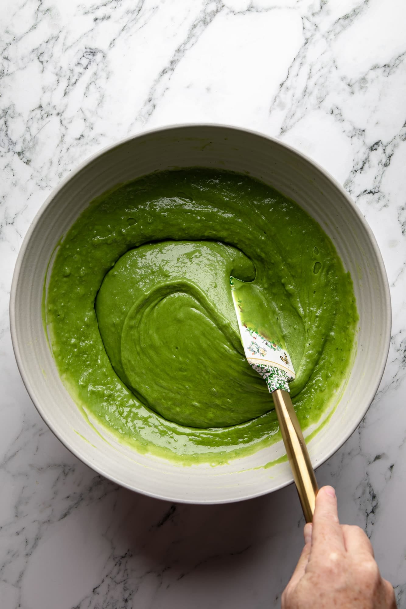 overhead view of a hand stirring the batter for vegan strawberry matcha cake with a spatula in a large bowl.