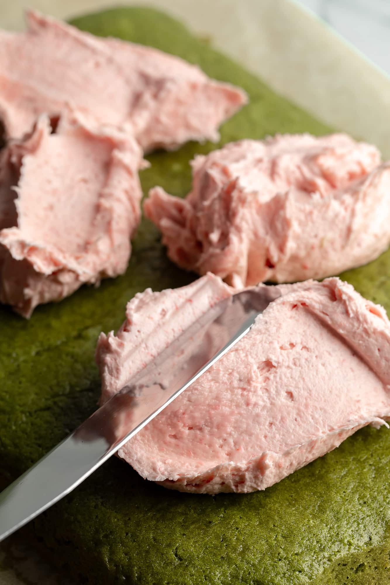 close up on a knife spreading strawberry frosting on top of a baked matcha cake.