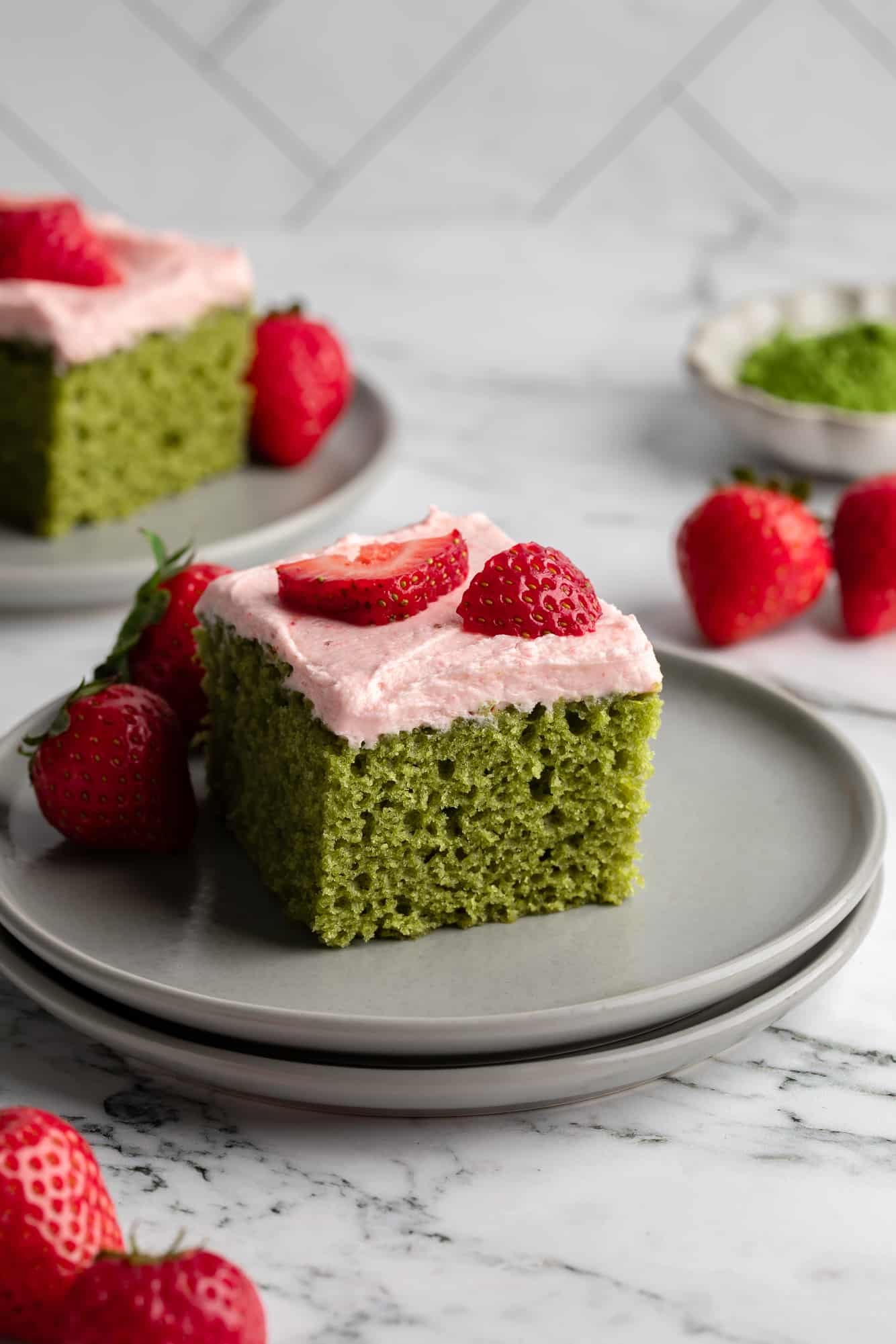 side view of a square slice of vegan strawberry matcha cake topped with fresh strawberries on a grey plate.