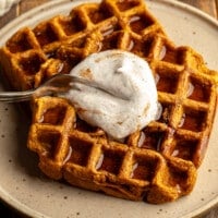 Close up on a fork cutting into vegan pumpkin waffles topped with vegan whipped cream on a plate.
