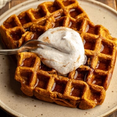 Close up on a fork cutting into vegan pumpkin waffles topped with vegan whipped cream on a plate.