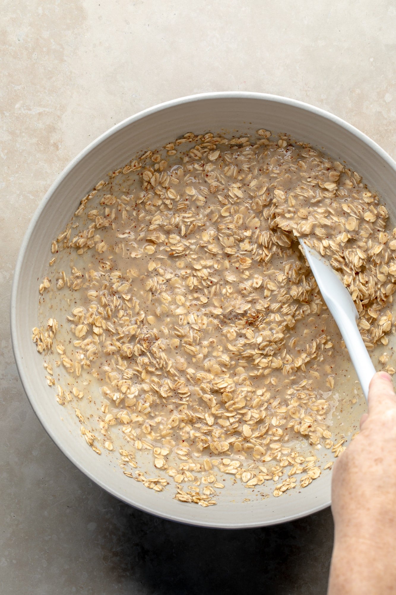 overhead view of a hand using a white spatula to stir the wet oatmeal mixture for baked berry oatmeal in a large bowl.