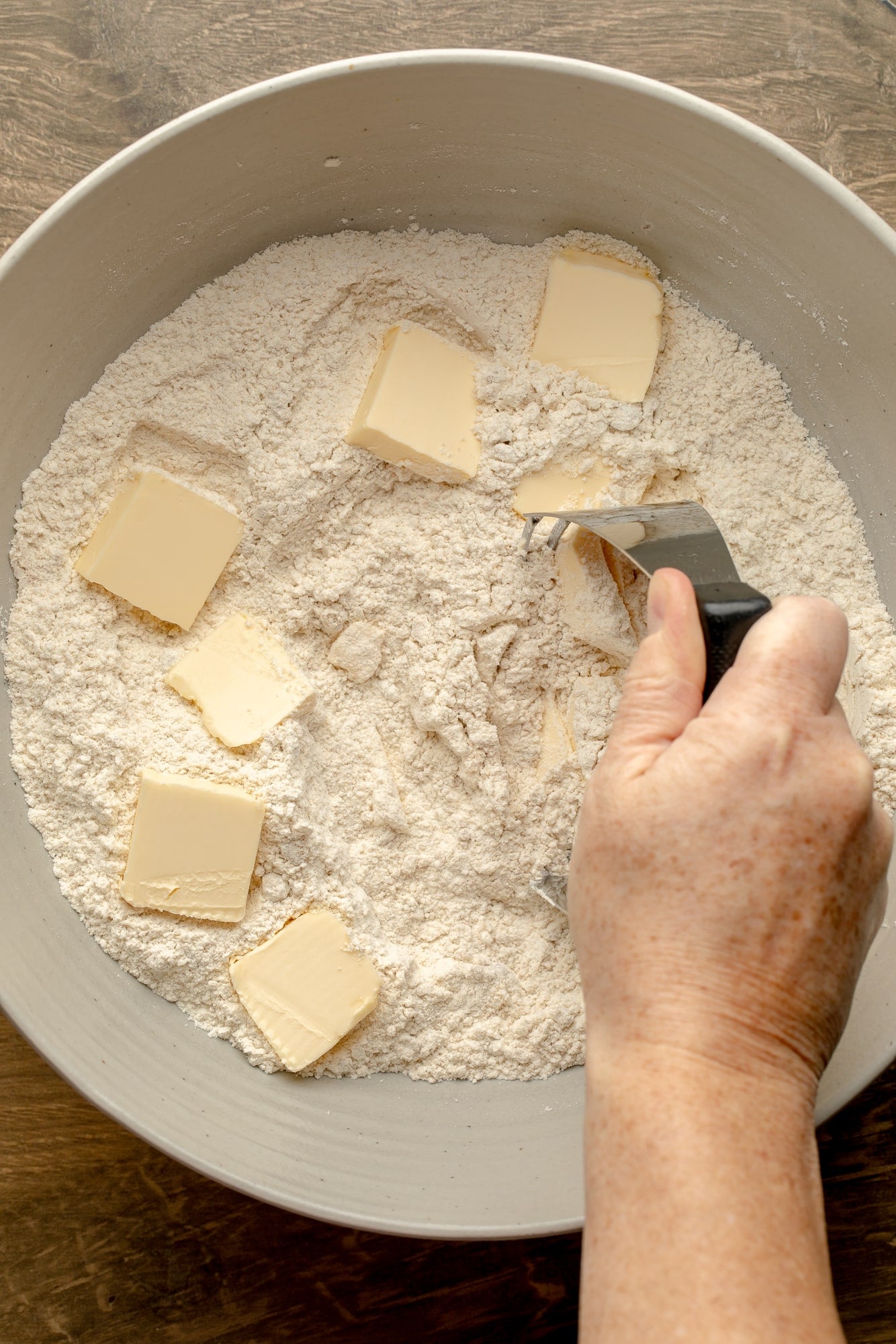 overhead view of a hand using a pastry cutter to work butter cubes into a dry flour mixture in a large bowl.