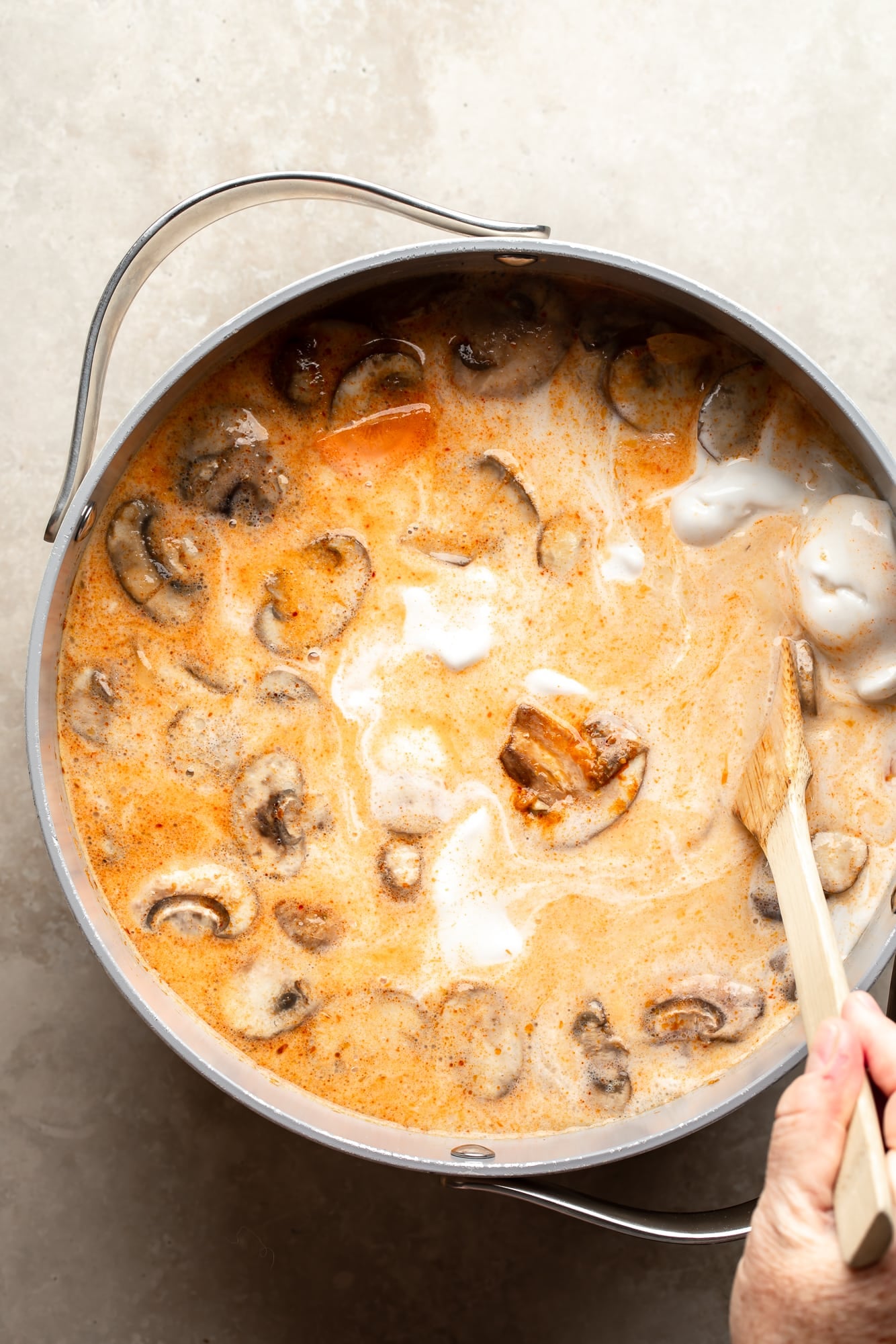 overhead view of a hand using a wooden spoon to stir Thai Coconut Soup (Vegan Tom Kha) in a large pot.