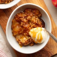 square image of a bowl of vegan apple crisp topped with caramel and a scoop of vanilla ice cream.