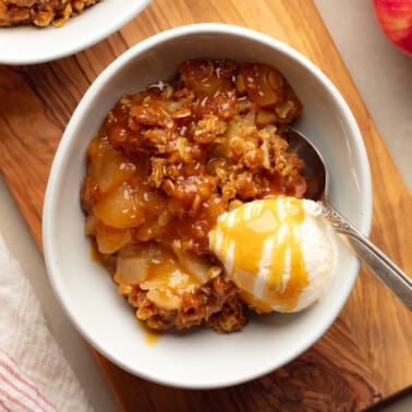 square image of a bowl of vegan apple crisp topped with caramel and a scoop of vanilla ice cream.